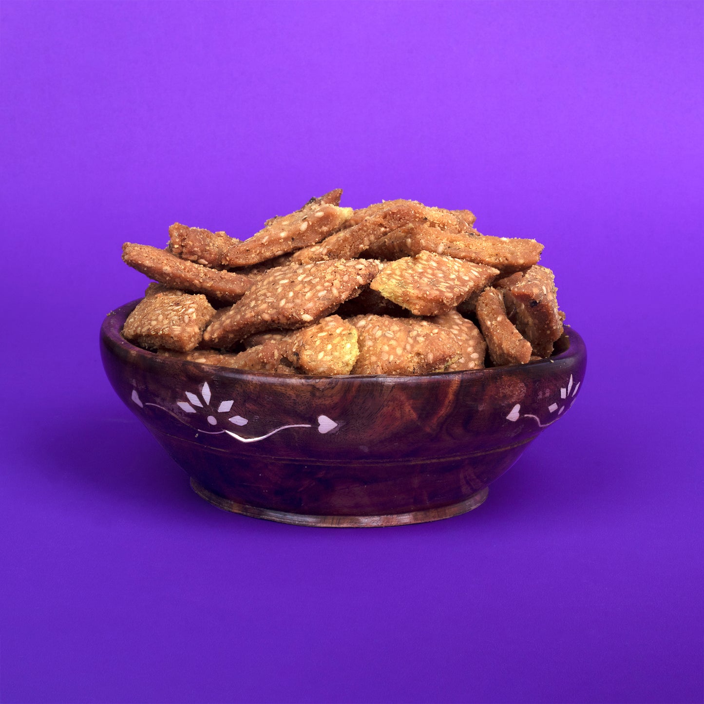 A decorative wooden bowl filled with golden-brown Til Sakkarpara, a traditional Gujarati sweet snack made from sesame seeds and jaggery, placed against a bright purple background.
