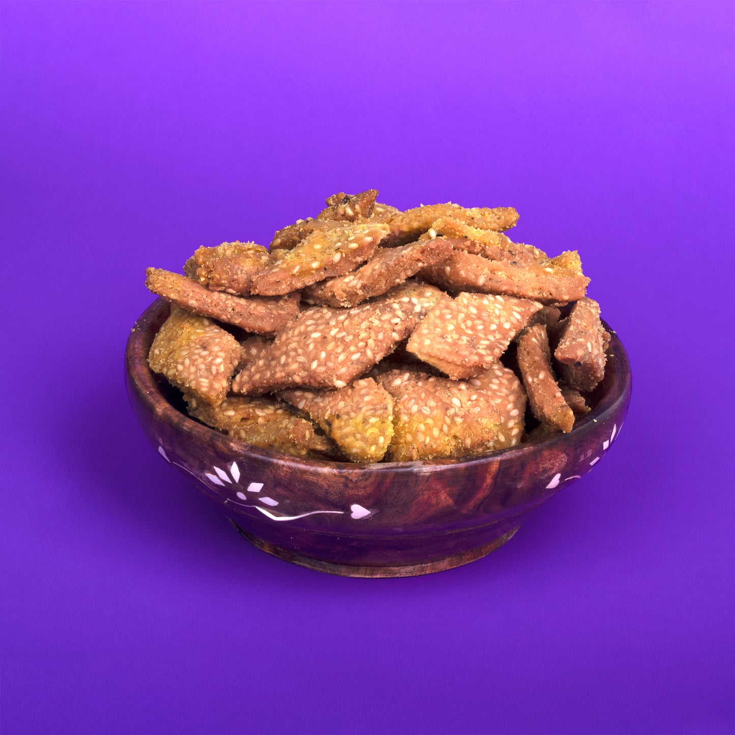 A decorative wooden bowl filled with golden-brown Til Sakkarpara, a traditional Gujarati sweet snack made from sesame seeds and jaggery, placed against a bright purple background.